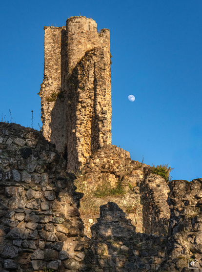 Color photo of a nearly full moon in a dark blue sky next to the ruins of a medieval castle tower lit by the setting sun. 