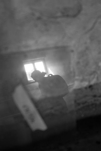 Im Vordergrund unscharf eine  Glasoberfläche mit Schmutz.  Dahinter als Silhouette eine Person mit Rucksack. Im Hintergrund ein helles, quadratisches Fenster, durch das starkes Licht hereinfällt und die Figur gegen das Licht stellt