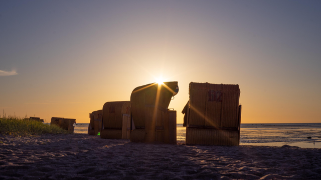 Farbfoto im Querformat. Zu sehen sind mehrere Strankörbe im Licht der untegehenden Sonne, im Hintergrund flaches Wasser, im Vordergrund Sand. Die Sonne bildet einen Stern mit sechs Zacken an der Ecke eines Strandkorbs. 