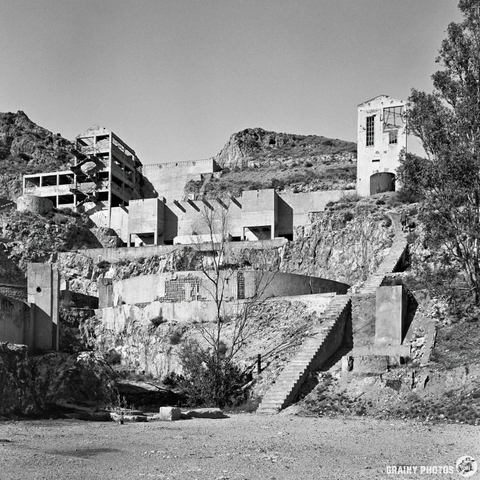 Abandoned concrete structures on rocky terrain, featuring crumbling buildings and staircases. The scene is in black and white, emphasizing the desolation and decay of the site surrounded by sparse vegetation and steep cliffs.