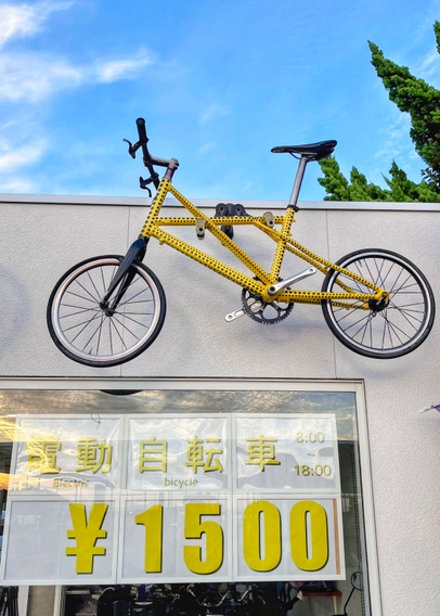 Ein kleines gelbes Fahrrad, mit kleinen schwarzen Punkten hängt als Eyecatcher an der Fassade eines Fahrradverleihs. Im Schaufenster ist in gelber Schrift der Preis von 1500 Yen zu lesen. Da Naoshima sehr hügelig ist, empfiehlt sich meistens doch ein ebenfalls günstiges E-Bike.