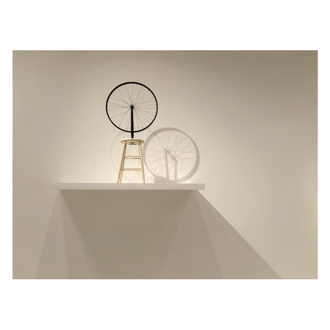 Photo of a bicycle wheel with its fork attached upside-down to a wooden stool, which stands on a shelf mounted high on a plain white wall. Lighting from above throws the wheel’s shadow onto the wall so that there appear to be two wheels oriented diagonally to each other. The shelf also casts its shadow below it in triangles of varying shades.

I took this photo at the Marcel Duchamp retrospective at the Museum of Modern Art in NYC. The original “Bicycle Wheel” has been lost and this appears to be one of Duchamp’s later versions, part of his series of “readymades” or found objects presented as art.