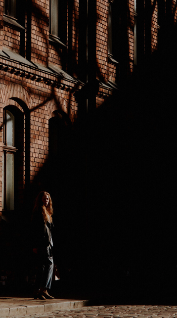 High contrast sunset backlit photo of a woman standing in front of red brick building covered by shadow.