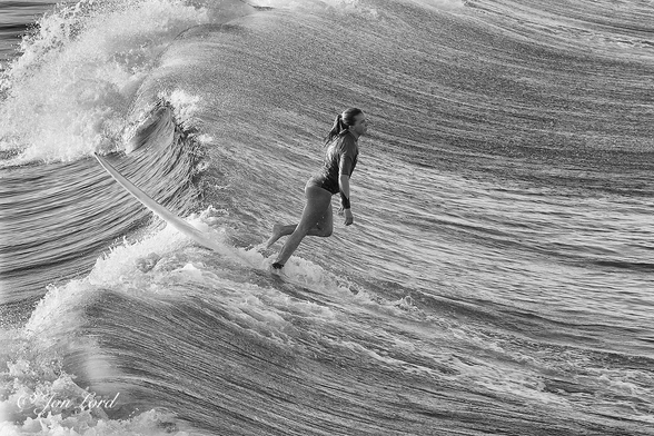 This is a black and white sports photo in landscape format of a young female surfer standing on her board behind a breaking wave. Hermosa Beach, California (2013).

To the right of centre is the Pacific Ocean close to the shore. The sea has multiple streaks extending at right angles to the waves. The water rises sharply towards the breaking wave which dominates the left side of the image. The wave is viewed along its length and with it extending away from the camera and it breaking from right to left. In the centre of the photo is a young female surfer, the surfboard being just behind the crest of the breaking wave and pointing towards the shore. She is calmly walking towards the back of the board and about to enter the sea. Our surfer is viewed in profile with her right side in view and with her filling half the height of the photo. She has long wet hair, tied with a bunch at the back of her head and is wearing a t-shirt and has bare legs. The California Current flows south from the colder waters of Canada and the Pacific Northwest, so the sea must have been warm, since most surfers would wear wetsuits (or she didn't mind the cold ?)