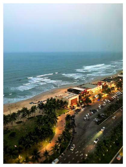 A high-altitude view of the beach at Đà Nẵng, Vietnam, facing the East Sea (or South China Sea) at sunset. A multi-lane avenue at bottom, dotted with cars with their headlights on, stretches alongside a wide pedestrian path illuminated by warm lights. The path in turn borders some low-rise buildings and a park lined with palm trees. Tiny figures of people walk everywhere. Beyond is a strip of beach and the blue-gray sea, with long lines of foam-edged waves rolling ashore. The distant horizon is softened by smog, and the featureless sky above glows faintly with the dying light. I took this photo through the window of a high-rise hotel room, and you can see some faint vertical streaks and smudges on the glass at right frame.