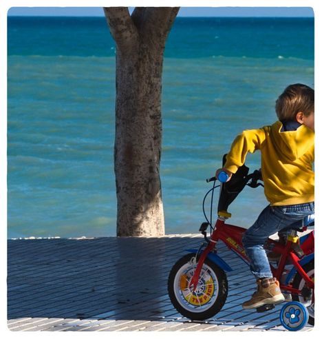 Auf einer Uferpromenade am Meer  fährt von links nach rechts ein nigelnagelneues rotes Kinderfahrrad mit Stützrädern vorbei, der Besitzer mit gelbem Hoodie blickt nach rechts aus dem Bildrahmen.