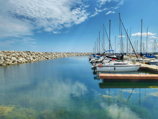 Sailboats are seen tied up to wooden docks at a marina. Calm water and a rock retaining wall is shown as well as clouds reflecting in the water.