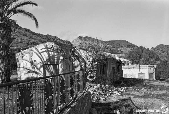 Abandoned buildings amidst desert terrain, with a palm tree nearby casting shadows. The structures show signs of decay, surrounded by rocky ground and sparse vegetation in a monochrome setting.