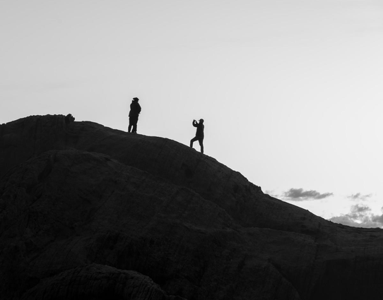 A black and white landscape photo of people standing on a nearby hill silhouetted against a bright sky. The hill rises from the lower right toward the upper left and then starts to curve down again. There's a couple of clouds in the distance in the lower right otherwise the sky is clear. The Hill is dark and no detail can be seen. One person is standing to the right and lower than the other two (which kind of looks like one person because they stand so close together). The person on the right is in a pose that appears to suggest they are taking a photo of the other two standing higher on the hill to the the left. 