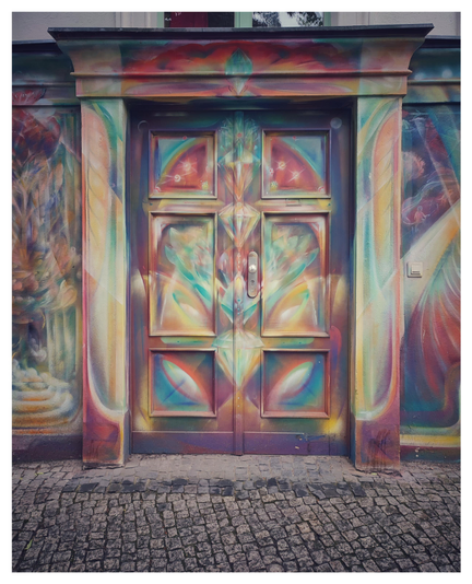 Farbfoto im Querformat. Zu sehen ist ein Teil einer Hauswand, der komplett mit einem Auftragsgraffiti bemalt ist. In der Mitte des Fotos sieht man eine ebenfalls bemalte Tür zum Souterrain, wie sie für Kreuzberger Altbauten typisch ist.