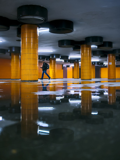 Person walking through an underpass with orange columns. Reflections of the columns and the person on a large puddle on the ground.
