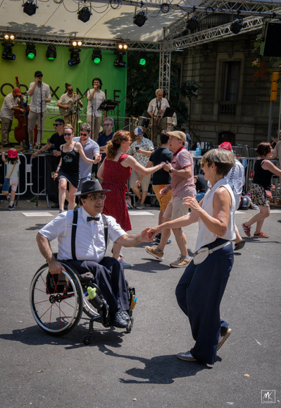 Color photo of a woman dancing with a man in a wheelchair in the middle of a street along with many other dancing couples and behind them is a stage with a jazz band playing music. 