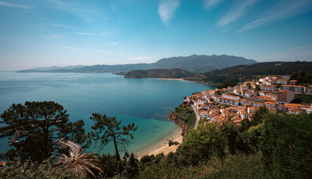 A view overlooking a scenic town with white walls and orange roofs above a lovely white sand beach with turquoise water. Green hills and cliffs in the foreground.