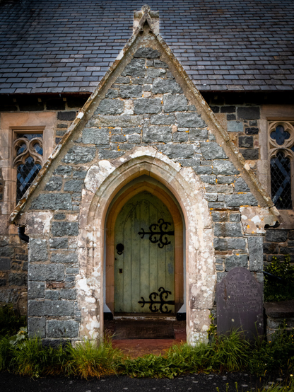 A stone entrance porch of a parish church in North Wales. The pointed archway leads to a pale green weathered wooden door made with vertical planks and curly decorative black iron hinges. To the right of the entrance an old slate gravestone stands partially hidden by the tall, overgrown grass in front of the abandoned church.