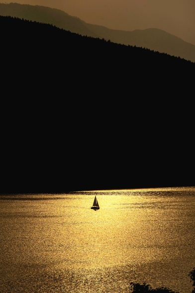 Das Foto zeigt ein einsames Segelboot, das bei Sonnenuntergang als dunkle Silhouette auf einem glitzernden See fährt. Das goldene Licht der tiefstehenden Sonne spiegelt sich intensiv auf der Wasseroberfläche und lässt diese fast wie flüssiges Gold leuchten. Über dem glänzenden Wasser erhebt sich eine massive, dunkle Bergkette, die fast den gesamten mittleren Teil des Bildes einnimmt und einen starken Kontrast zum hellen Wasser bildet. Im Hintergrund sind weitere Bergumrisse in einem sanften, dunstigen Braunton unter einem warmen, gedämpften Abendhimmel zu erkennen. Die gesamte Szene strahlt eine tiefe Ruhe und Einsamkeit aus.