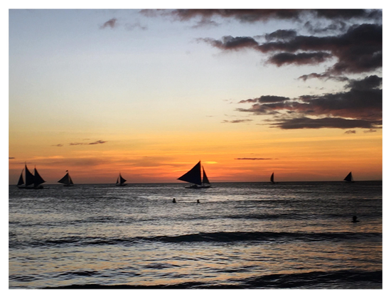 Photo on the same beach a couple of hours later, at sunset. The sky is a faded blue, transitioning to orange and gold at the horizon. Dark fingers of clouds push in from right frame. Silhouettes of sailboats with their unfurled triangular sails line the horizon, along with the dots of some distant swimmers.