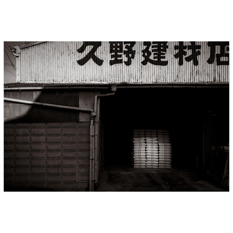 A black-and-white view of an industrial warehouse facade with corrugated metal siding and large painted Japanese characters across the upper wall. The open entrance leads into deep shadow, where a neatly stacked pile of sacks becomes visible only after looking closely. A glass block wall and angled pipe structure occupy the left side, while dim light reflects faintly across the concrete floor.