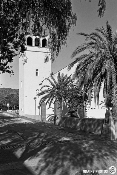 A black and white image of a building featuring a tower, surrounded by palm trees, with a clear view of the street leading up to it. The architecture suggests a quaint, possibly historic location.