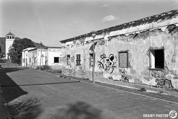 A deserted street featuring rundown buildings with graffiti, including a prominent spider design, under a clear sky. In the background, a tall structure resembling a tower adds to the scene's urban decay. The image is in black and white, emphasising textures