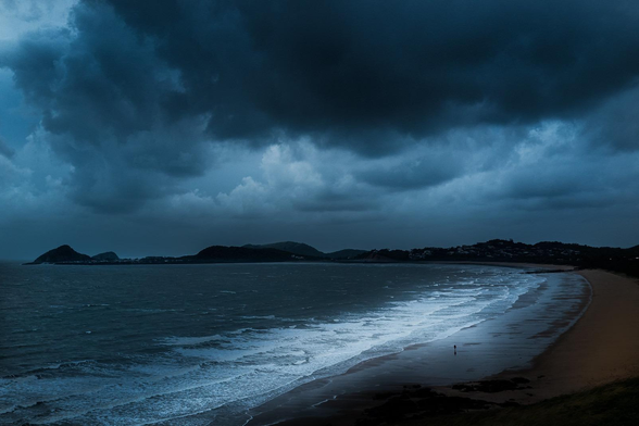 un homme minuscule marchant sur une plage déserte entre la mer et le ciel du même bleu sombre menaçant