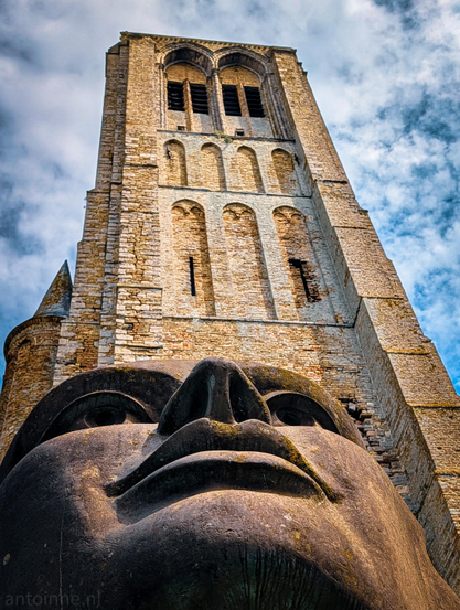 A massive, weathered stone sculpture of a human face looking upward, set against the backdrop of a tall, historic brick church tower.

The face is positioned at the very bottom of the frame, shot from an extreme close-up, low-angle perspective. It depicts a stylized human face from the chin up, highlighting thick lips, a broad nose, wide eyes, and a prominent brow.

Rising directly behind the sculpture is a massive, square medieval tower built from light-colored brick and stone. It features classic Gothic architectural elements, including arched alcoves, narrow slit windows, and decorative brickwork.