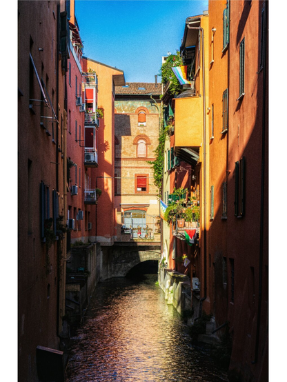 This photograph is of a narrow, picturesque canal and is flowing between tall, tightly packed residential buildings. The buildings have warm coloured vibrant facades in orange, terracotta, and yellow. They also have windows, small balconies, and window-mounted air conditioning units. (Lucky ducks) The sunlight is mostly down the centre of the passage photo and is illuminating the textured brick building at the far end and reflects across the surface of the water. There are several flags, including a rainbow flag and a Palestinian flag which are hanging from the balconies on the right side
 (💪🏻). In the background there is a small arched bridge which crossed over the water, where a few people can be seen walking behind the metal railing under the bright blue sky. This is il Canale delle Moline.