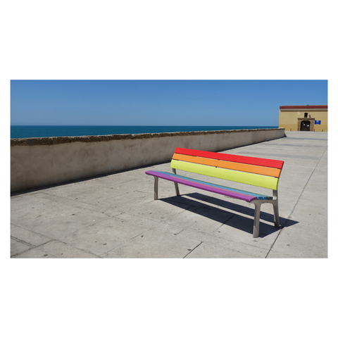 This is a landscape format colour image of a bench painted in rainbow colours sitting by the sea wall in Cadiz. The bench sits at an angle to the bottom right of the frame, while the sea wall crosses at an angle starting at the bottom left. The sea on the horizon creates a horizontal line crossing the frame. The sky is clear blue.