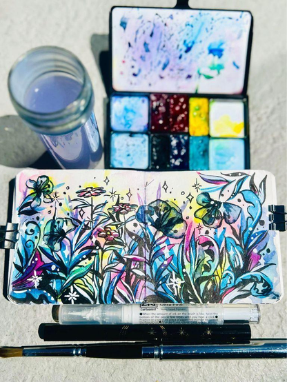 My watercolor and ink painting of a floral filigree flower garden in a sketchbook, surrounded by my artists’s gardening tools.