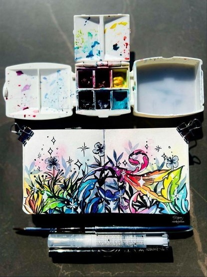 My watercolor and ink painting of flowing, colorful and rainbow florals & vines in a sketchbook. Also in the photo are my painting palette, watercolor paint, and paint brush and ink brush pens.