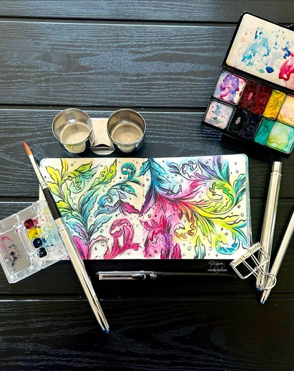 My watercolor and ink painting of a colorful flower and plant garden sparkling and glistening in the sun in my sketchbook. Also in the photo are my paint palette, rinsing water, and black and white inks.