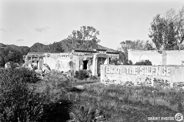 A black-and-white image of a dilapidated building surrounded by overgrown grass and shrubs. The structure features crumbling walls and a large painted message that reads "ENJOY THE SILENCE." Mountains are faintly visible in the background.