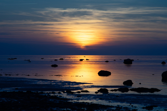 Farbfoto im Querformat. Zu sehen ist der Sonnenuntergang über der Ostsee bei wolkigem Himmel. Die Sonnenscheibe ist als Umriss dicht über dem Wasser zu sehen, ihr Licht spiegelt sich im Wasser.