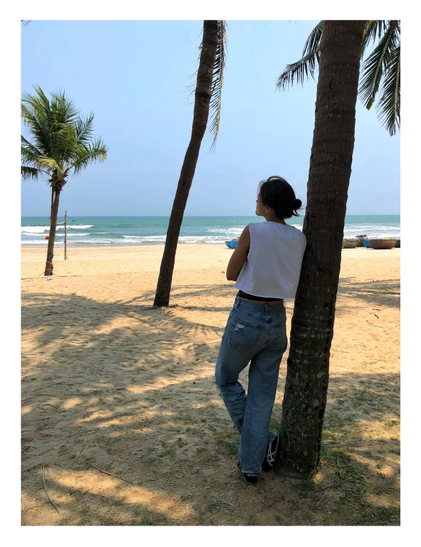 A wide beach with palm trees and a view of the distant strip of ocean under a clear sky. Shadows of palm leaves on the warm brown sand. A line of boats sits at the surf line. A person in a white sleeveless T-shirt and jeans, with their hair gathered in a bun, leans against the nearest tree trunk and looks out to the sea.