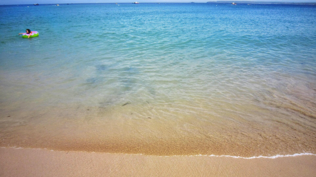 Fine golden sand at St Ives beach gently slopes into crystal-clear turquoise water with beautiful colour gradients from light aquamarine to deeper blue. A person relaxes on a circular green and pink inflatable ring in the very shallow, calm sea. Gentle waves lap at the shore, and the water's exceptional transparency reveals the light brown sandy seabed beneath. In the distance, a few small boats dot the horizon under a clear blue sky, capturing the peaceful, tranquil beauty of this iconic Cornish coastal location.