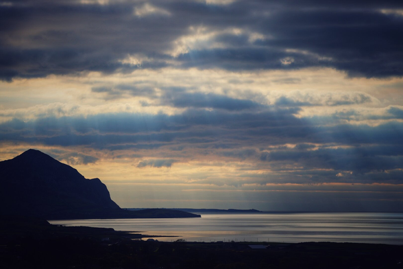 Dramatic landscape at dusk featuring a dark, jagged mountain silhouette on the left. The sky is filled with thick, layered grey clouds illuminated by golden sunlight breaking through. Below, a calm body of water reflects the subtle light, with a distant peninsula visible on the right horizon. The foreground is shadowed, creating stark contrast with the luminous sky.