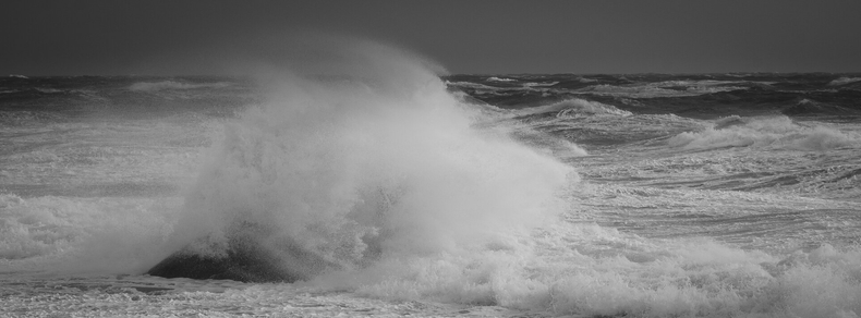 A monochrome panoramic photograph of a coastal sea on a stormy day. The storm blows from right to left, creating large foamy waves on the sea. Slightly to the left in the image is a sizeable black rock in the sea, almost completely obscured by foam and spray from a wave that has broken against it. The wind is clearly present in how it drags the spray and foam with it.

The image is quite bright from a light cloud cover overhead, with the sunlight coming from top right, while the horizon is quite dark.