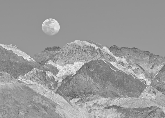 A black and white landscape photo of a full Moon rising over a barren rocky mountain peak. The mountain peak is composed of many different layers of rock which in color are many different colors, but in monochrome it is many shades of gray. The layers are uneven, bent, and jagged. The Moon is rising over the left side of the image.