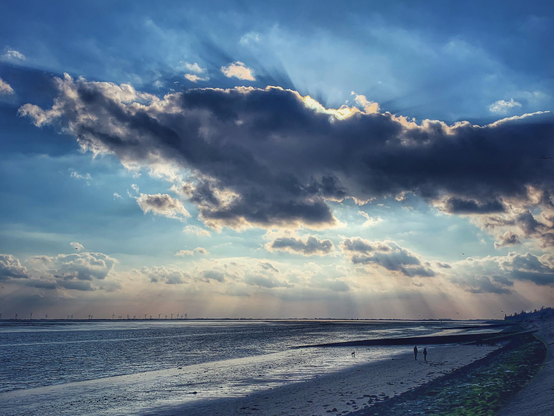 Sonnenuntergang hinter dunkelgrauen Wolken an der Südstrand-Promenade in Wilhelmshaven. Die Sonne wirft sichtbare Strahlen nach oben und unten und erzeugt dadurch eine mystische Stimmung im Wattenmeer. 