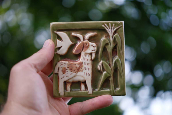 a hand holding a ceramic tile. the design is a goat admiring a corn plant. a white bird is flying in the sky behind the goat.