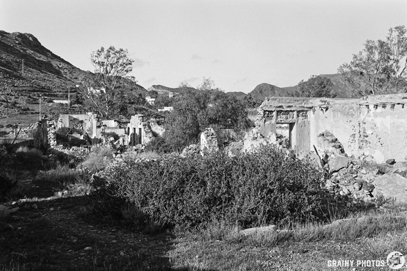 A desolate landscape featuring the ruins of building structures, scattered debris, and overgrown vegetation. The scene is set against a backdrop of hills, conveying a sense of abandonment and nature reclaiming the area.