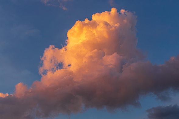 Photograph of a large cumulus cloud in a blue sky. It is late in the day, and the setting sun lends yellow, orange, and purple tints to the various portions of the fluffy cloud that is highlighted by sunlight or in shadow. The sky is dark blue due to the fading light.