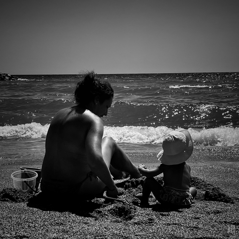 En la imagen en blanco y negro se ve a una mujer adulta sentada en la arena junto a una niña pequeña con sombrero, frente al mar. Están cerca de la orilla, con pequeñas olas rompiendo y un cubo de juguete al lado, transmitiendo un momento tranquilo y familiar en la playa.