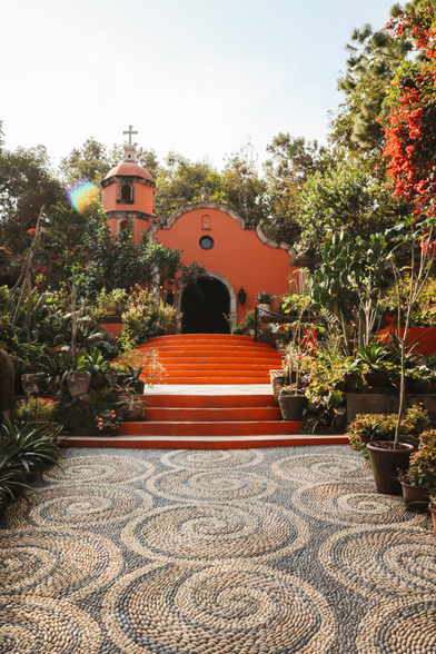 A vibrant terracotta-orange chapel sits nestled among lush green tropical trees and flowering plants under a bright, clear sky. In the foreground, a wide pathway features an intricate, mesmerizing mosaic pattern made of small river pebbles arranged in large, interlocking spirals. The pathway leads to a set of wide, terracotta-colored concrete steps that ascend toward the arched entryway of the chapel. Potted plants and cacti line the steps and walkways.