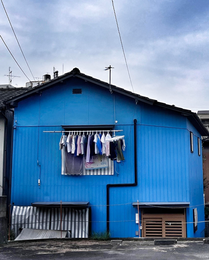 Ein blaues Holz-Haus bei bewölktem Himmel. Auf Höhe des einzigen Fensters ist eine Stange befestigt, an der Wäsche hängt, hauptsächlich T-Shirts und Sweatshirts. Rechts an der Stange hängt ein kleines Extragitter für kleinere Teile. 