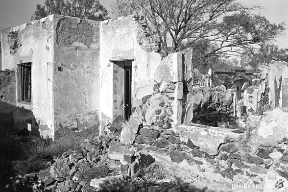 A dilapidated structure with crumbling walls and stones scattered around, surrounded by nature. The building's remains suggest it has been abandoned for a long time, with trees in the background adding to the sombre atmosphere.