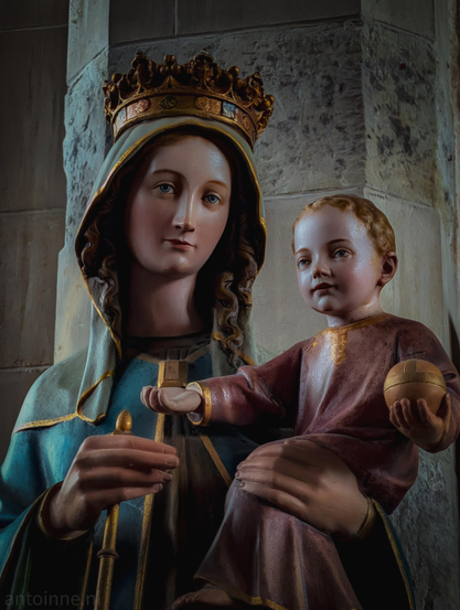 A sculpture depicting the Virgin Mary holding the infant Jesus. 

She wears an ornate, golden crown detailed with small geometric patterns over a light-colored veil or hood that frames her face. Curled brown hair peeks out from beneath the veil.

He is held on the right side of the frame, seated on Mary's arm and facing forward. He is dressed in a simple, reddish-brown tunic with gold trim along the neckline and cuffs.

The figures are set against a rustic, light-colored stone pillar. The texture of the stone is rough and weathered.