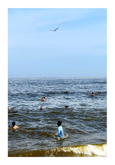Ocean waves dotted with swimmers under a blue sky lightly streaked with high clouds. Foaming waves break on the shore at the bottom of the photo. A swimmer wearing a long-sleeved blue shirt stands waist-deep in the water, vertically aligned with a seagull flying overhead in the distance.

Taken on a sunny day at Sandy Hook Beach in New Jersey.