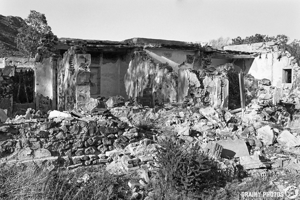 A black and white photograph of a dilapidated building surrounded by rubble and overgrown vegetation, highlighting the decay and abandonment of the structure amidst a desolate landscape.