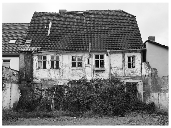 Schwarz-Weiß-Foto im Querformat mit weißem Rand. Zu sehen ist ein altes, mit Pflanzen teilweise überwuchertes Fachwerkhaus zwischen neueren Häusern.