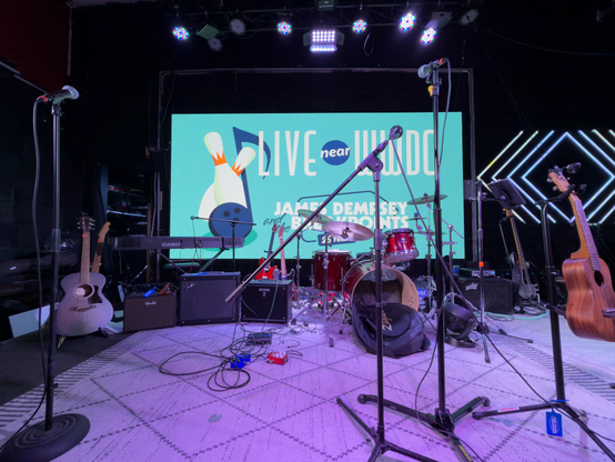 A purple-lit stage with band equipment on it. A large screen in the background shows a cyan poster that says “Live near WWDC”.